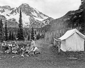 Camping party of men and women cooking at campfire and eating near tent in Indian Henry, Mt. Rainier National Park, Washington,